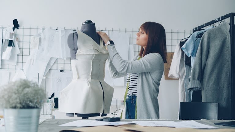 Fashion designer adjusting a dress on a mannequin in a studio filled with sketches.